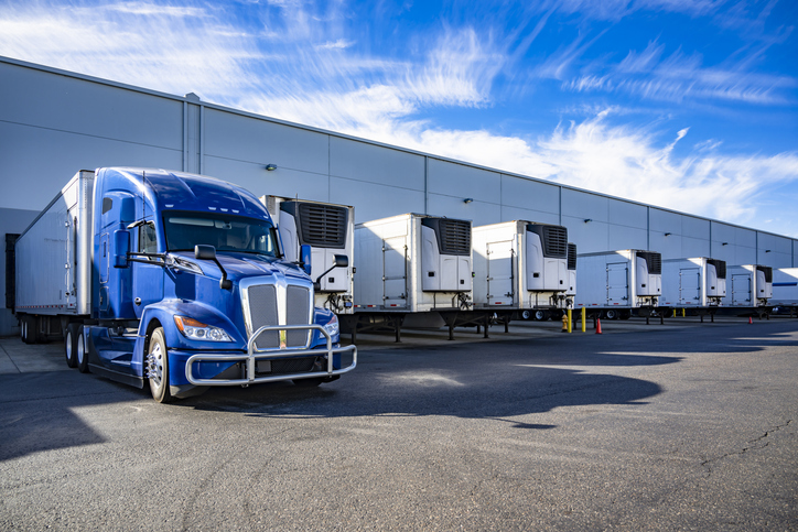 Blue big rig semi truck with extended cab for truck driver rest and refrigerator semi trailer standing in row with another semi trailers in warehouse dock gates loading cargo for next freight